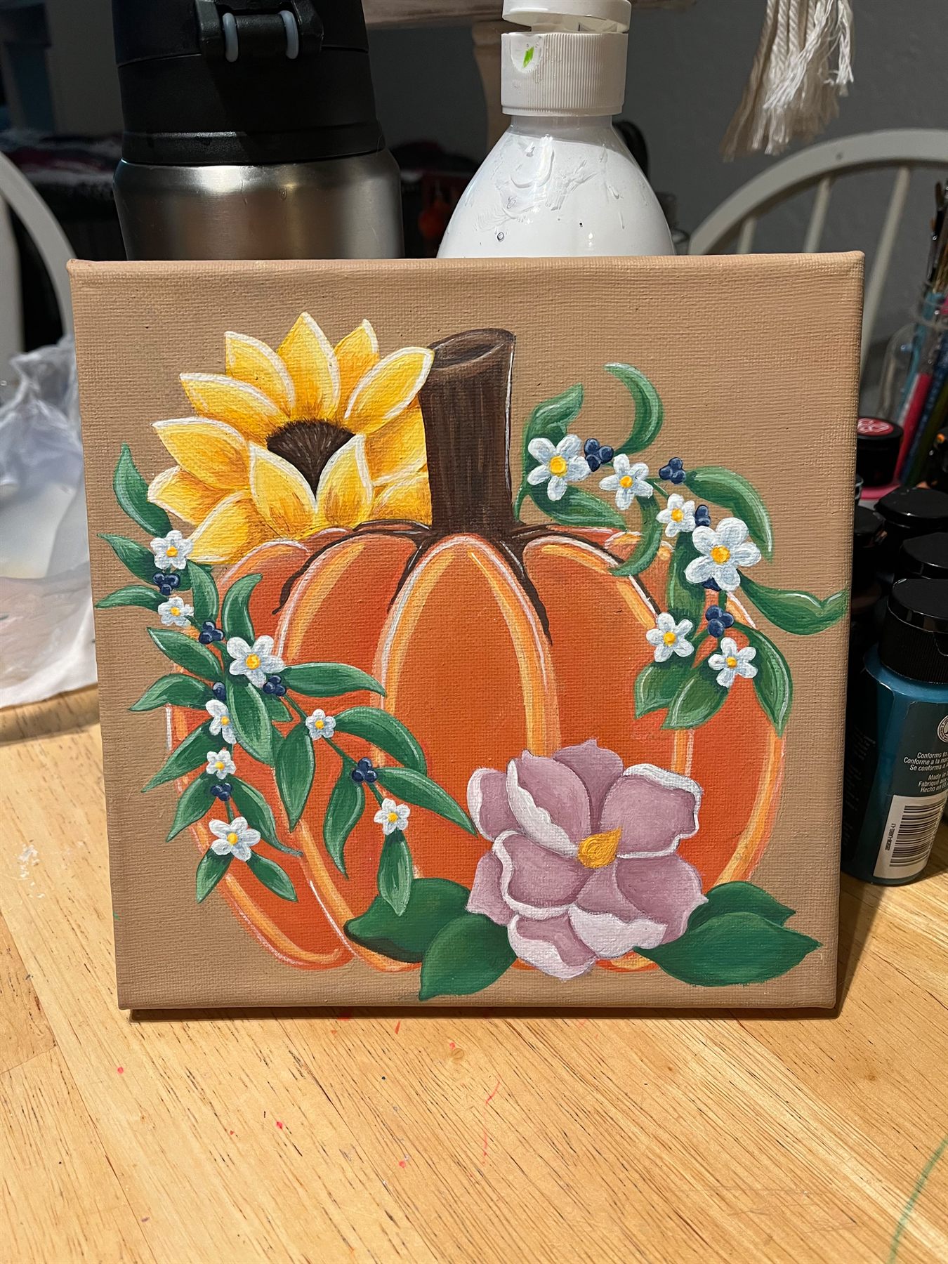 Painting of an orange pumpkin framed by a yellow sunflower, white blossoms, blue berries, and a lavender magnolia on a warm tan canvas