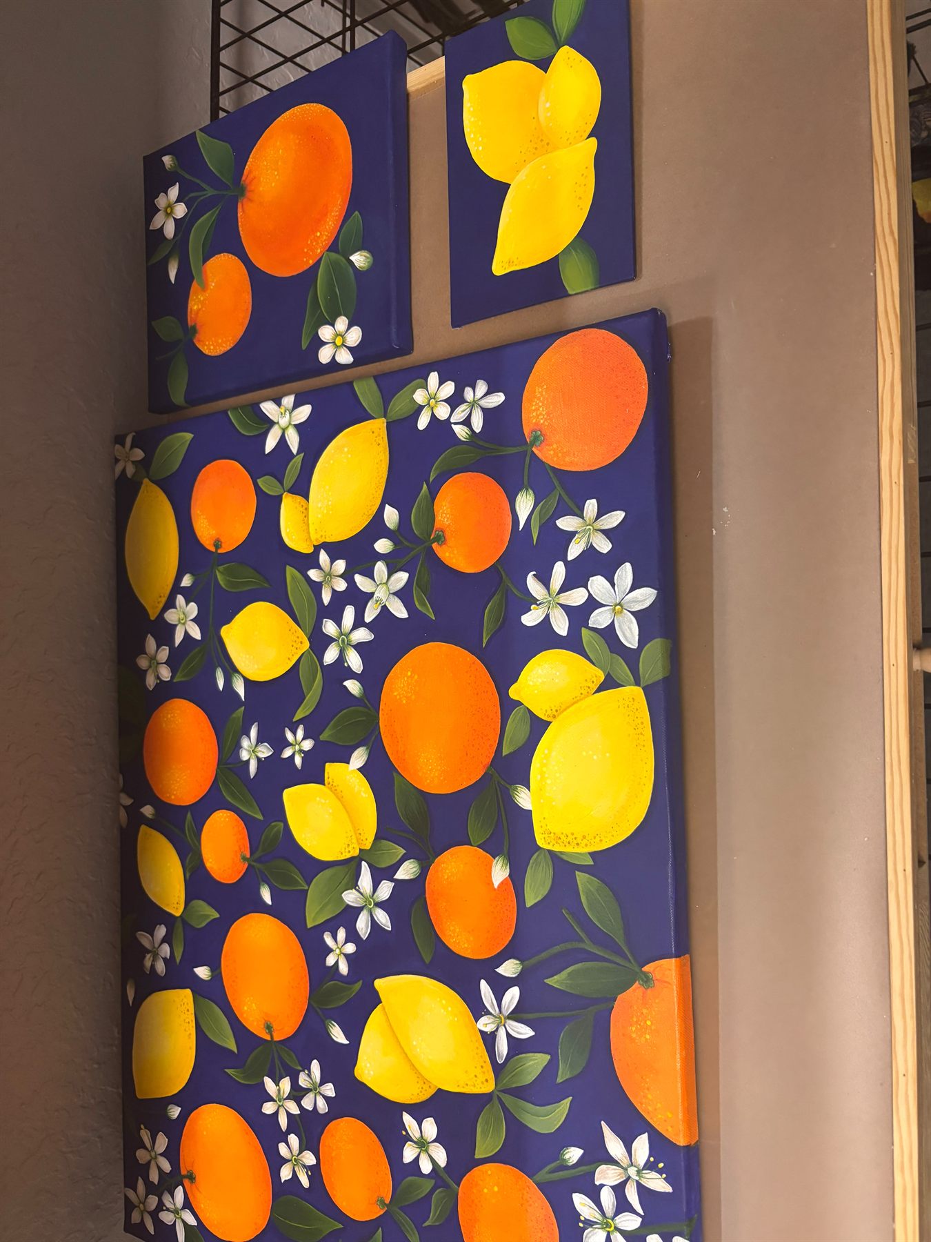 Three of Jenifer Mazer's navy citrus paintings hung together on a wall — two small canvases of oranges and lemons above a large citrus pattern canvas