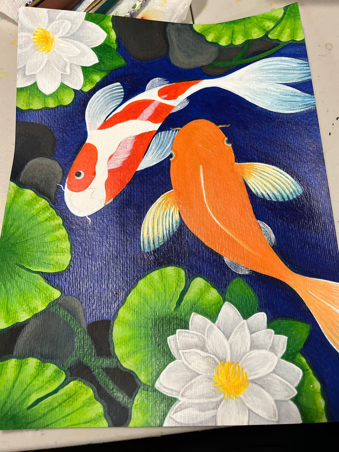 Painting of two koi fish, one orange and one red and white, swimming among green lily pads and white water lilies on deep blue water