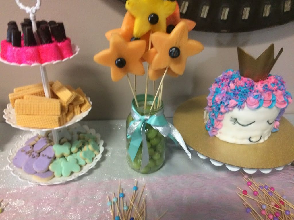 Styled shower dessert table with a tiered stand of cookies and wafers, melon stars in a jar, and a small cake with a gold crown and pink and blue ruffled frosting