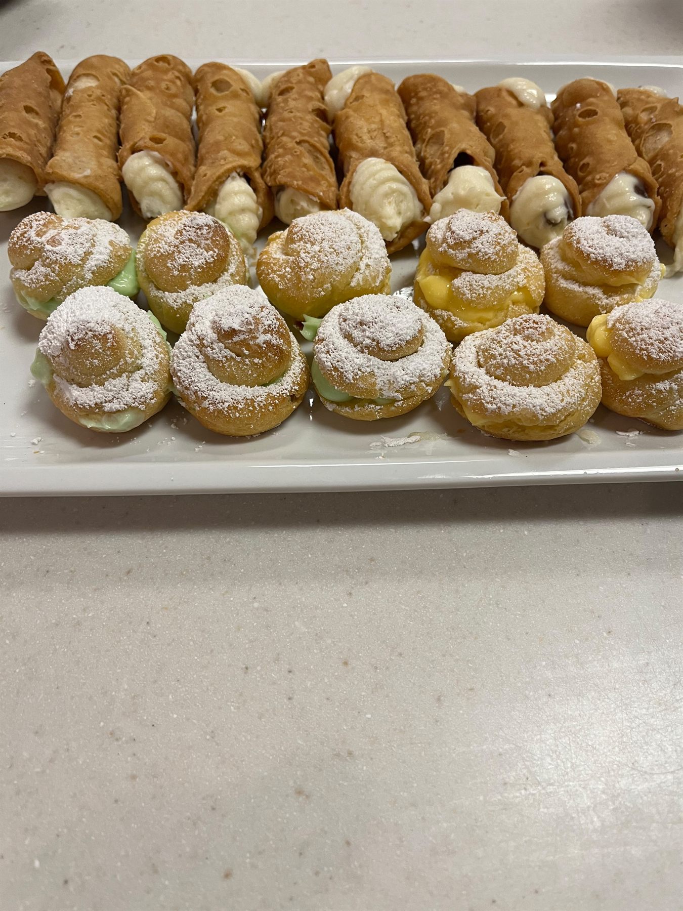 Platter of filled cannoli behind powdered-sugar cream puffs with pistachio, vanilla and lemon fillings