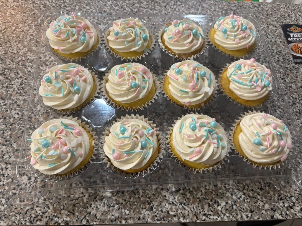 Gender reveal cupcake cut in half showing a bright blue centre inside vanilla cake, in front of a box of frosted cupcakes with black and silver sprinkles