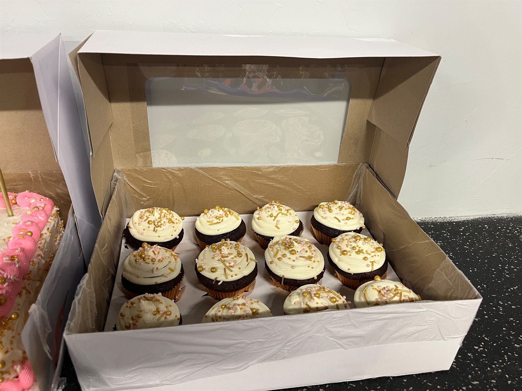 Bakery box of chocolate cupcakes with gold and blush sprinkles beside a pink and gold decorated cake, packed for delivery