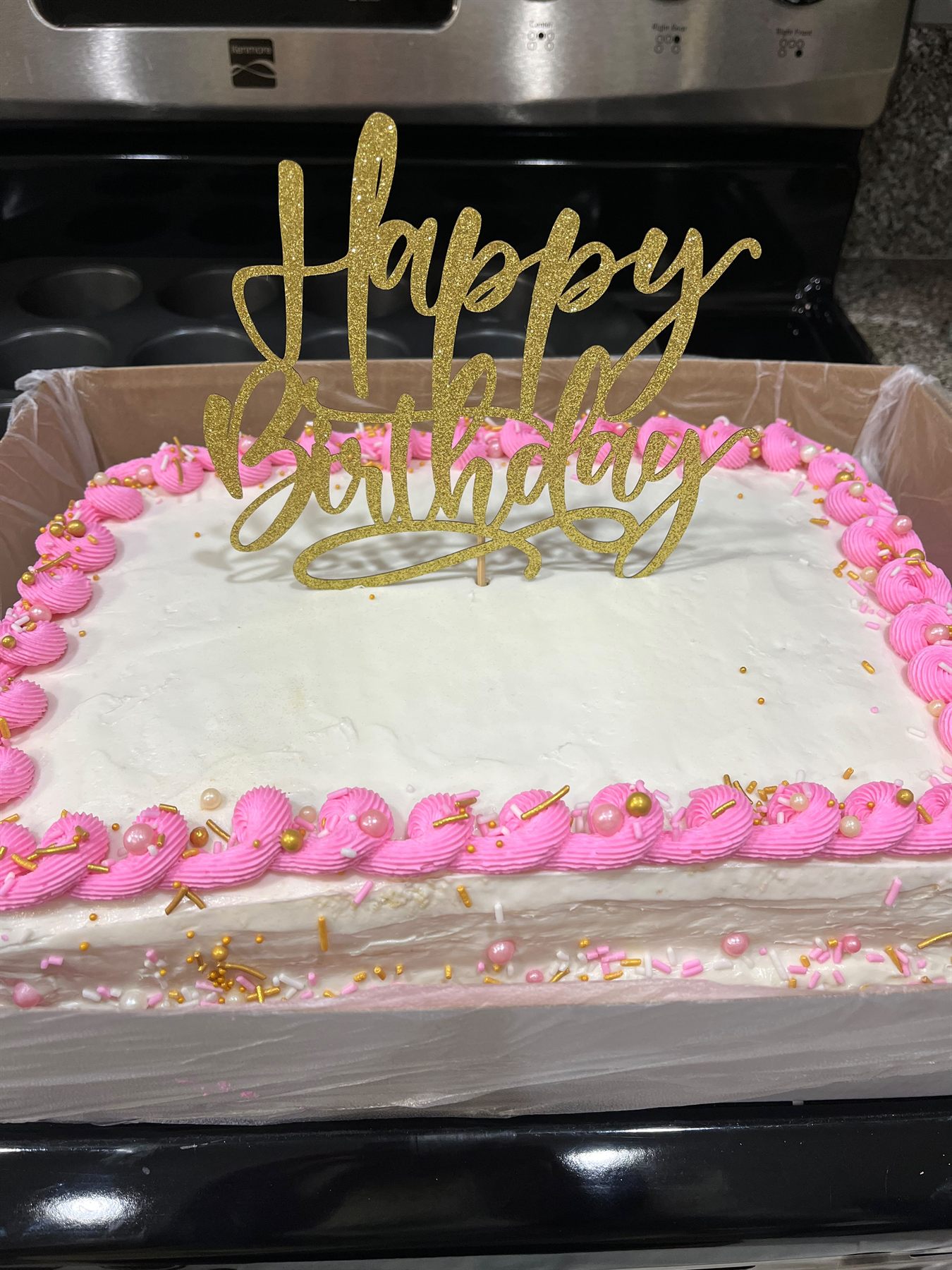 White buttercream sheet cake with pink piped shell border, gold and pink sprinkles, and a gold glitter Happy Birthday topper