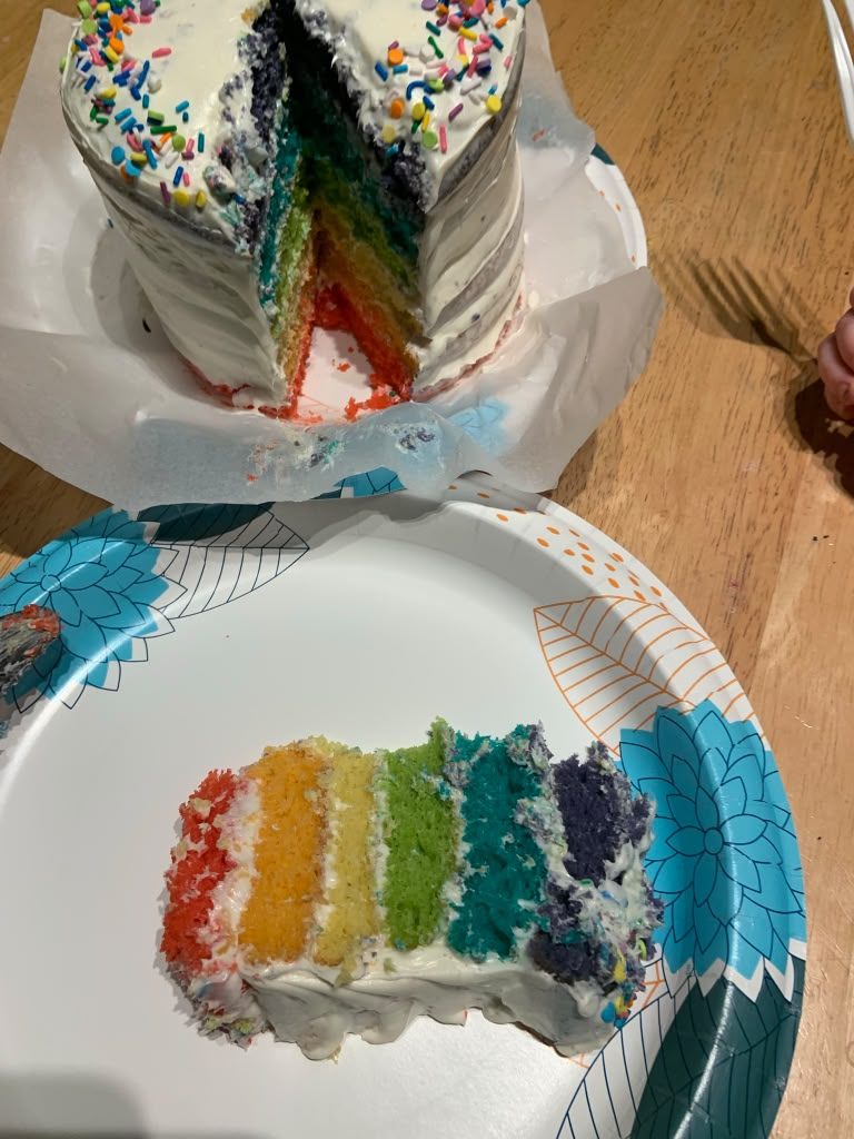 Cut rainbow layer cake showing six coloured sponge layers under white frosting and rainbow sprinkles, with a slice on a plate