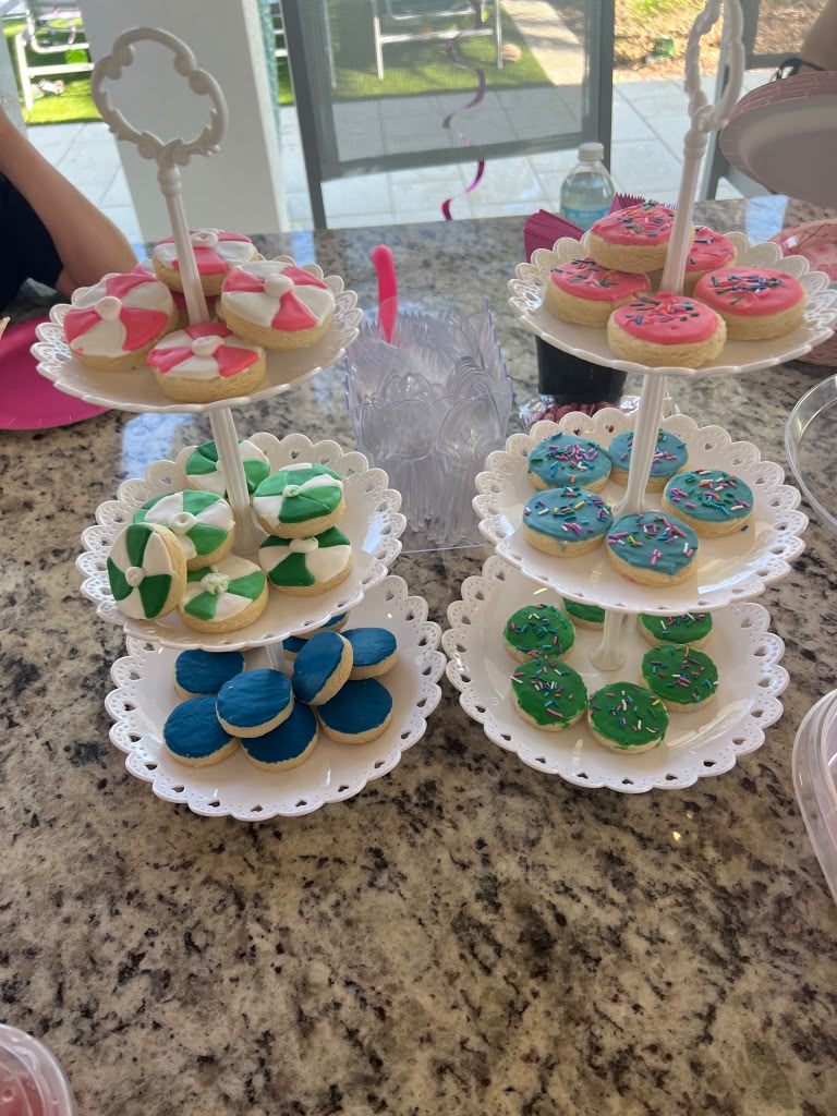 Two white tiered dessert stands holding iced cookies in pink, green, blue and sprinkle-topped designs