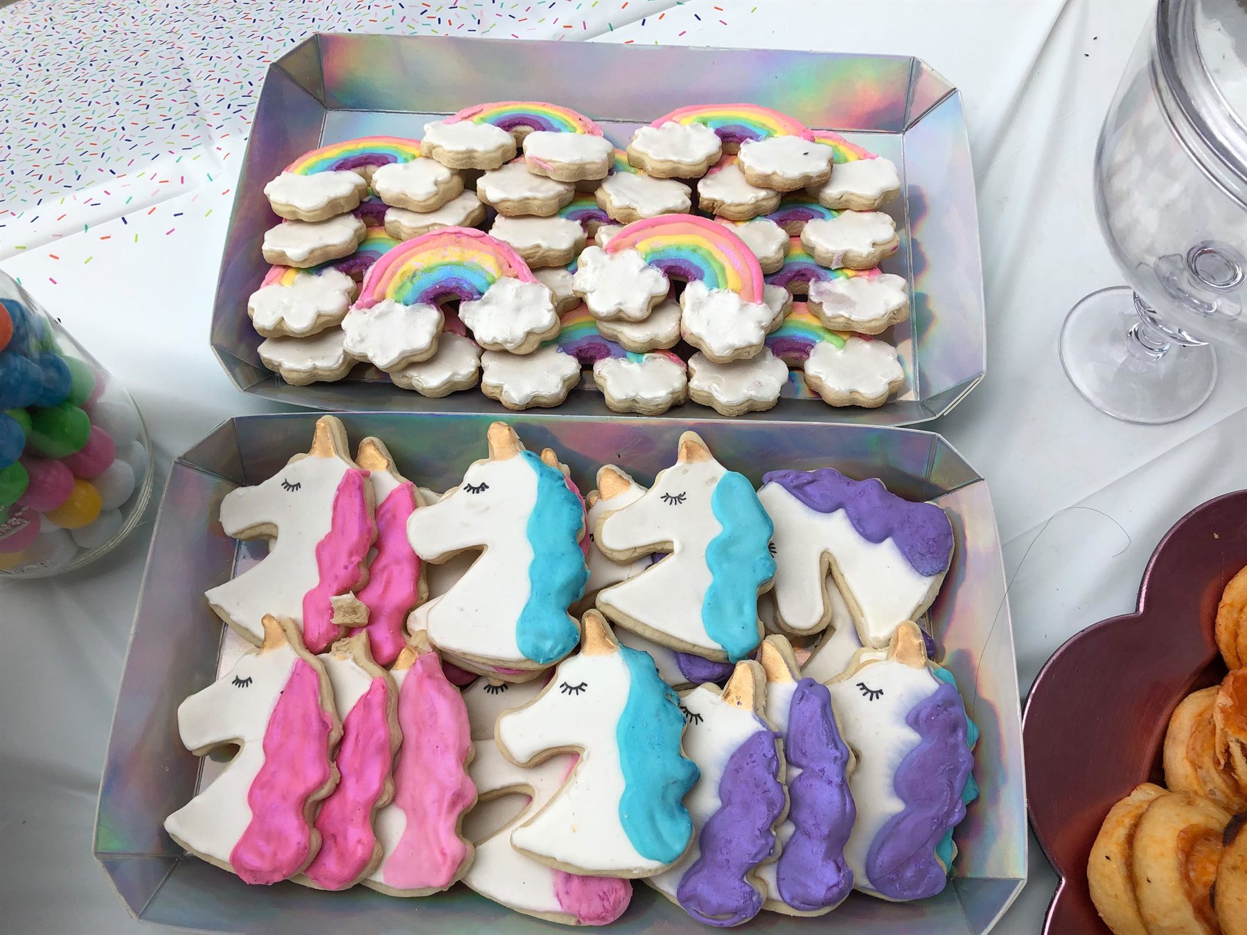 Iridescent trays of pastel rainbow and cloud sugar cookies above pink, blue and purple unicorn head cookies with gold horns